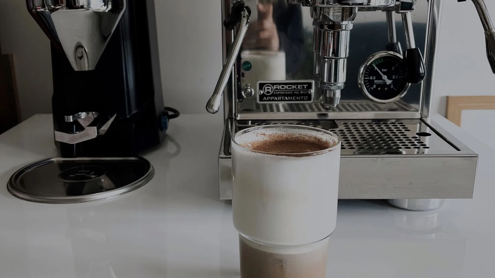 Photo by Feyza Yaşar on Pexels A white cup of coffee in front of a stainless steel espresso machine and grinder