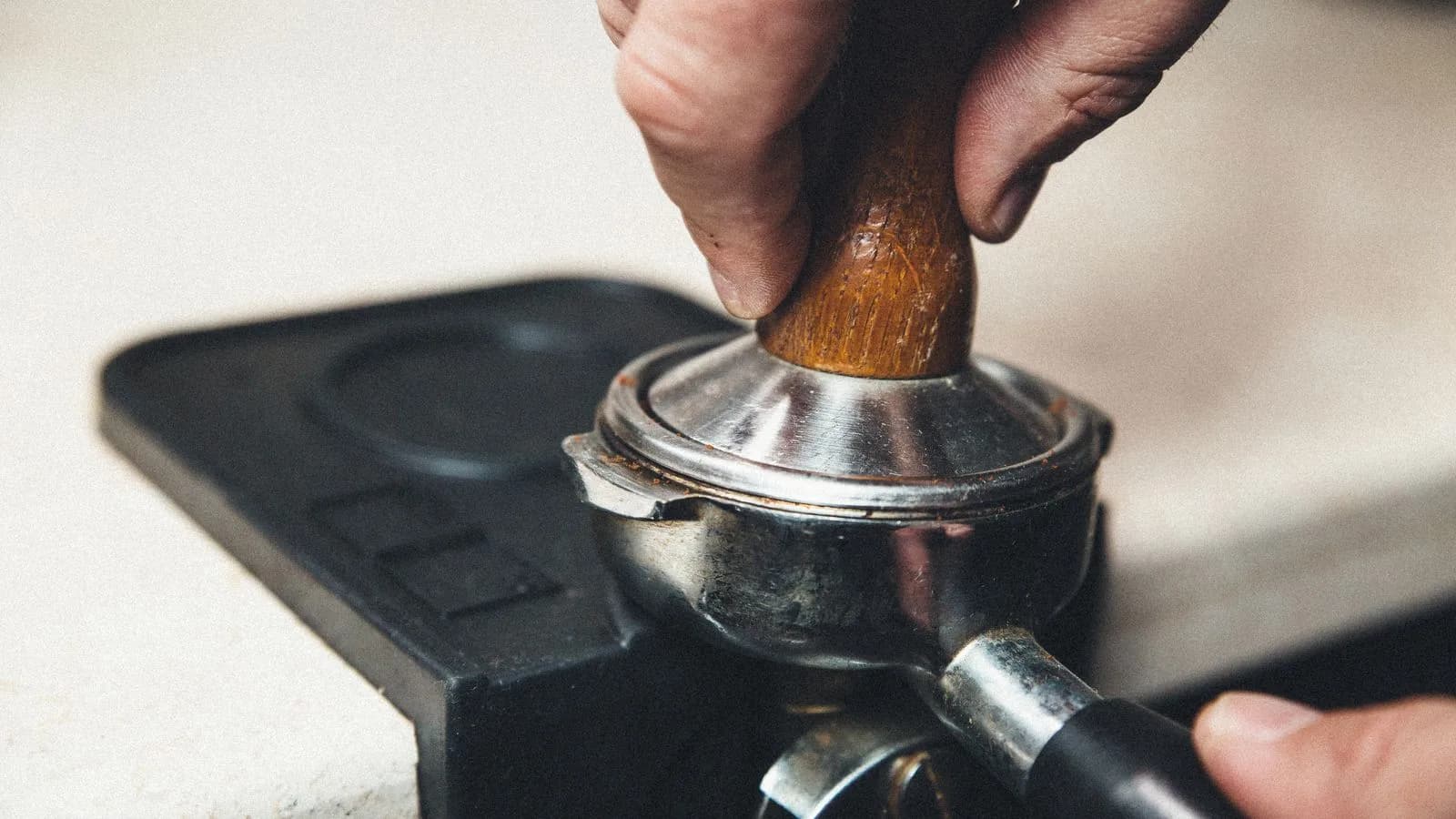 Photo by Claro Cafe on Pexels Fingers pressing a wooden-handled tamper down into a steel portafilter basket that rests on the edge of a black tamping mat